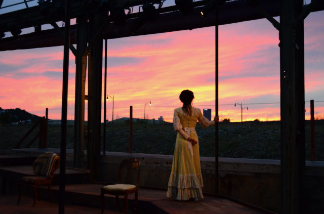 A woman in an old-style dress looks out at the sunset from within a steel structure.