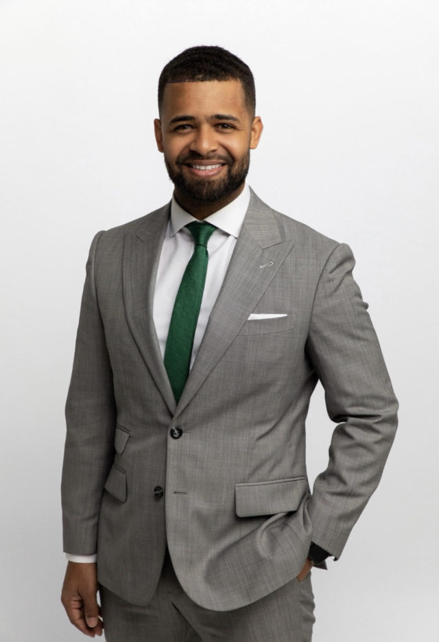 Justin Binion has a neat beard and hair and wears a slim grey suit with green tie.