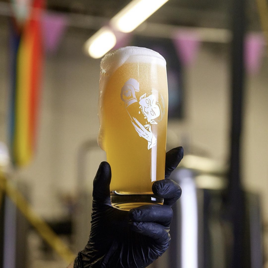 A gloved hand holds a beer up near a Pride flag.
